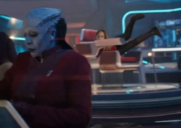 Lura Thok in the foreground, with Captain Ake in the background, peeking over the armrest of her Captain's chair, with her legs nearly straight up in the air
