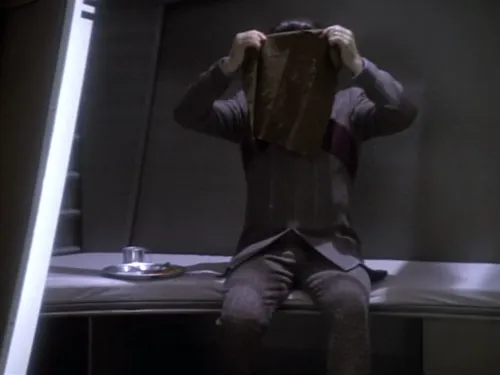A Cardassian in a holding cell holding a cloth over his face