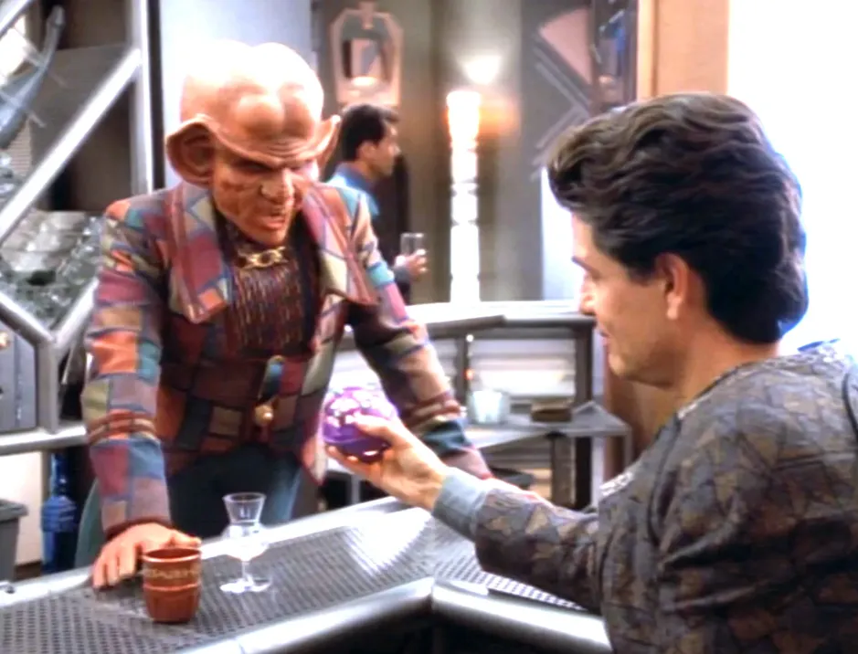 A man showing Quark a strange hand-held device from across the bar