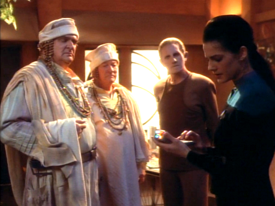 Odo and Dax standing with two men dressed in beige robes and beads