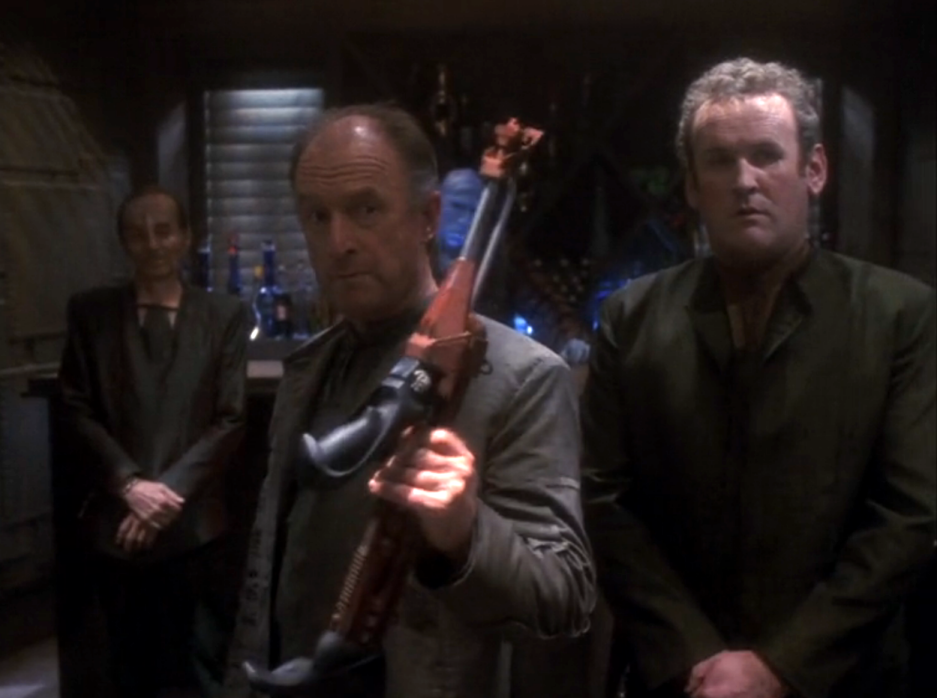 O'Brien in civilian clothing watching as another man holds a Klingon rifle