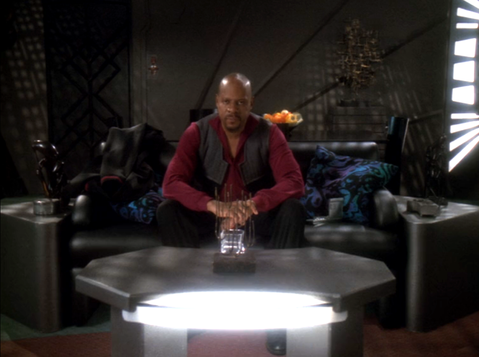 Sisko sitting on a couch, looking sternly directly into the camera