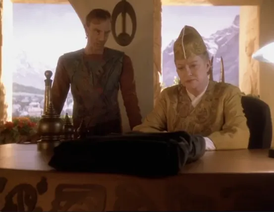 Kai Winn looking concerned at a book wrapped in black cloth on her desk, as Dukat watches over her shoulder