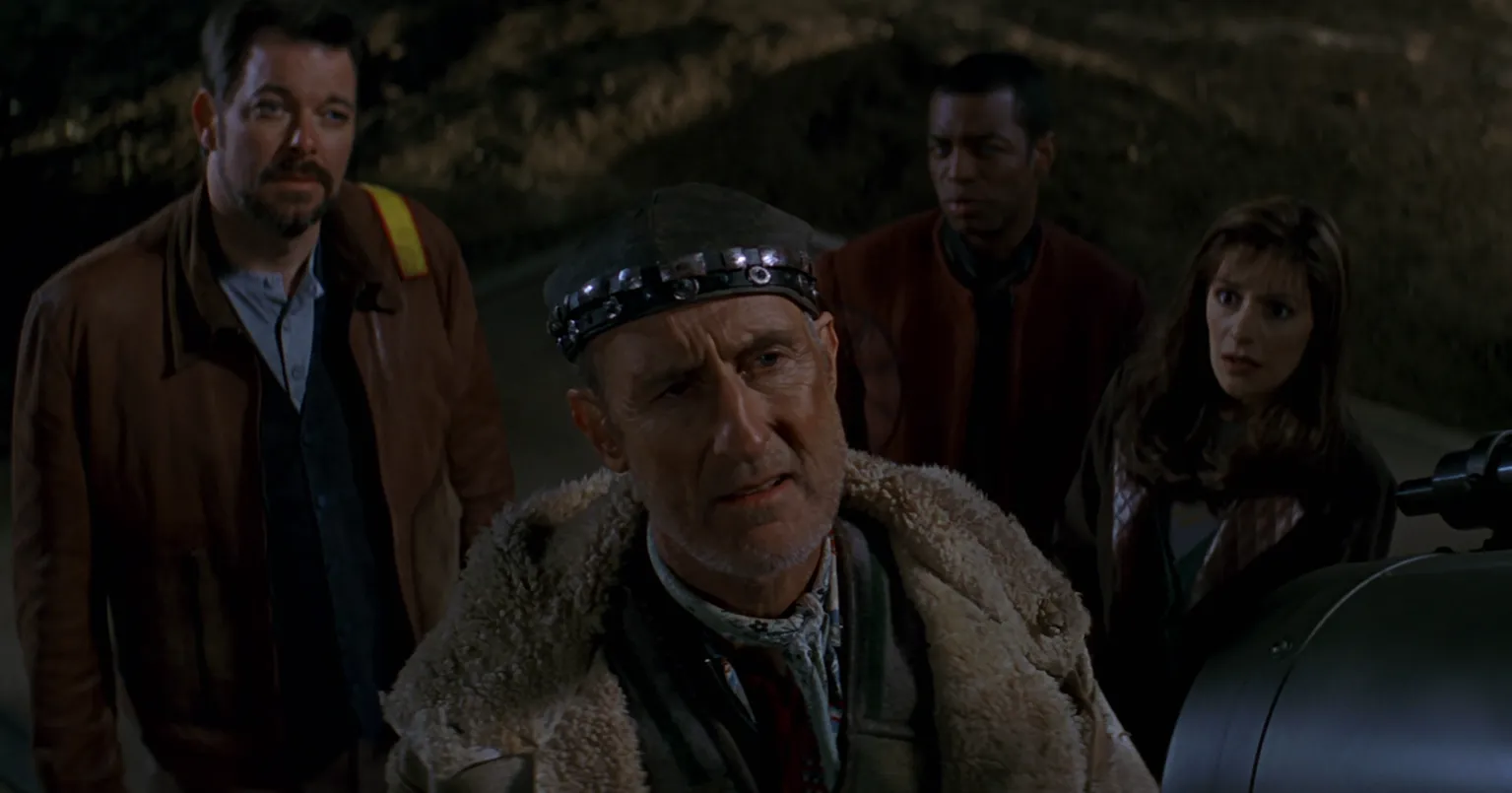 Cochrane looking pensively into the night sky as Riker, La Forge, and Troi look on