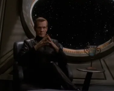 Luther Sloan sitting in a chair, legs crossed, looking pensive 