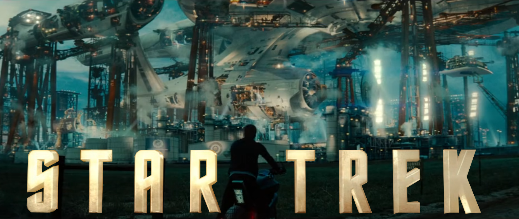 Kirk, resting on his motorcycle, looking up at a large Starfleet ship being built on the surface