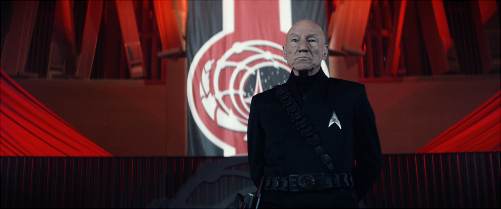 Picard, wearing all black and with a strange combadge, standing on a stage lined with red banners