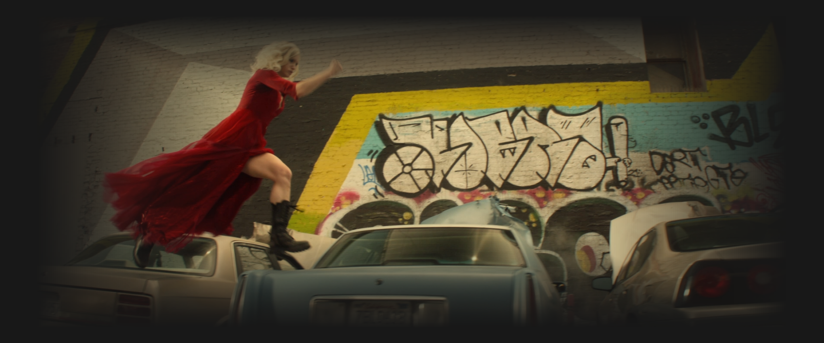 Jurati running atop several parked cars in a tattered red dress and black boots
