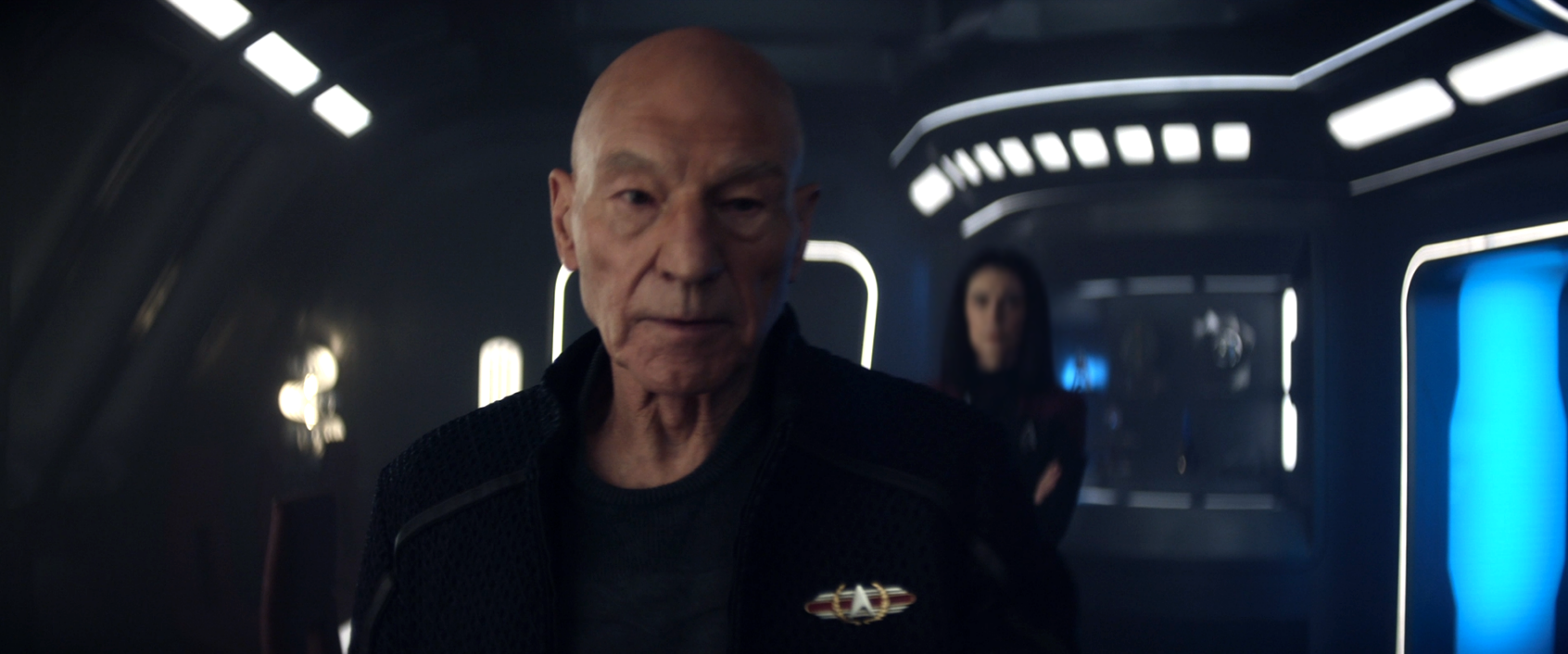 An unknown woman with her arms folded looking over Picard's shoulder