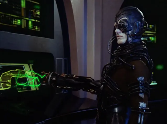 A pale humanoid covered in tubes and technology, extending a device on his arm toward a computer display, with the device emitting green energy pulses