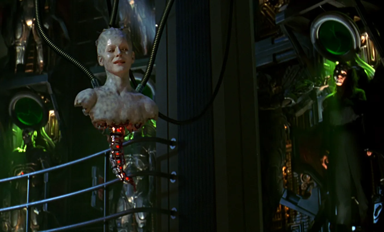 The head and shoulders of the Borg Queen being lowered from the ceiling by electronic hoses, with Borg in alcoves in the background