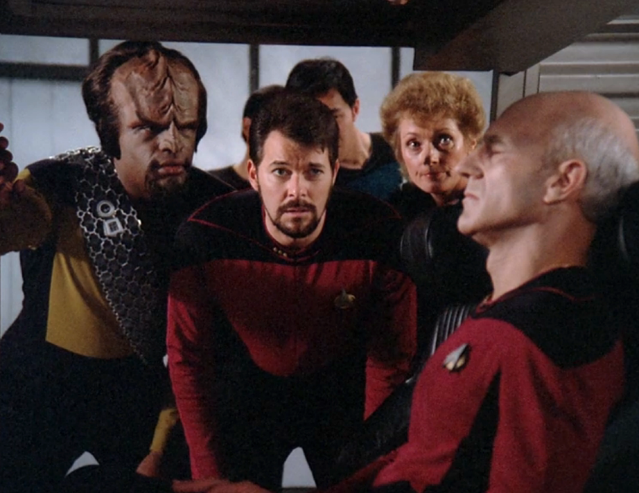 Worf, Riker, and Pulaski observing an unconscious Picard in a shuttlecraft