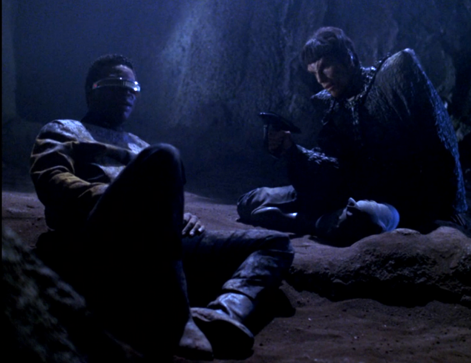 A Romulan holding La Forget at gunpoint in a dark, dusty cave