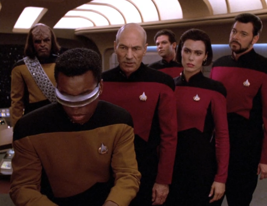 Most of the bridge crew and one unidentified man observing a console at the rear of the bridge