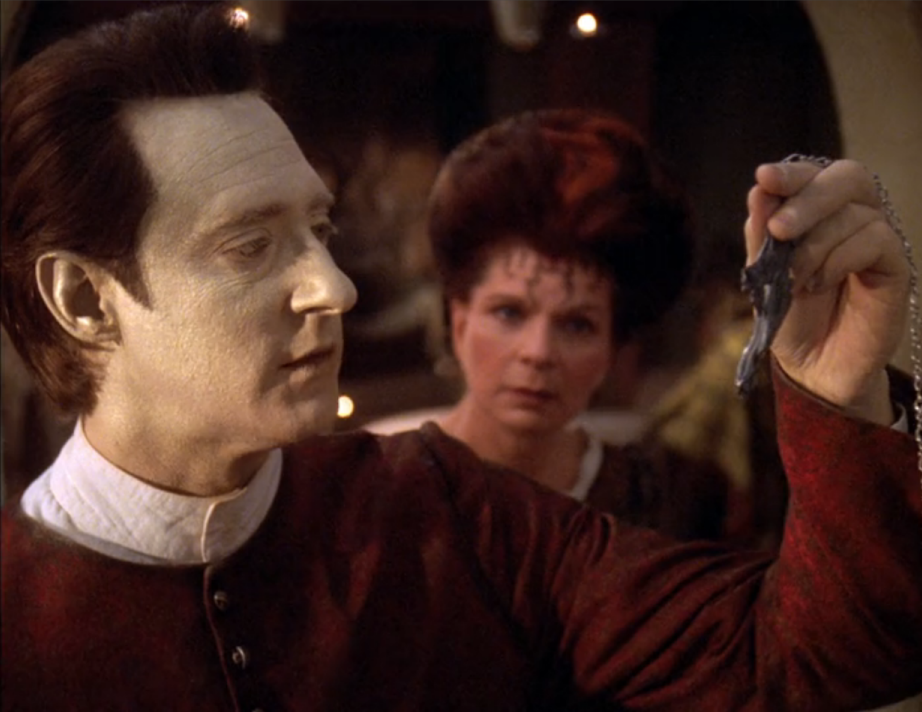 Data examining a strange piece of metal as Talur watches him