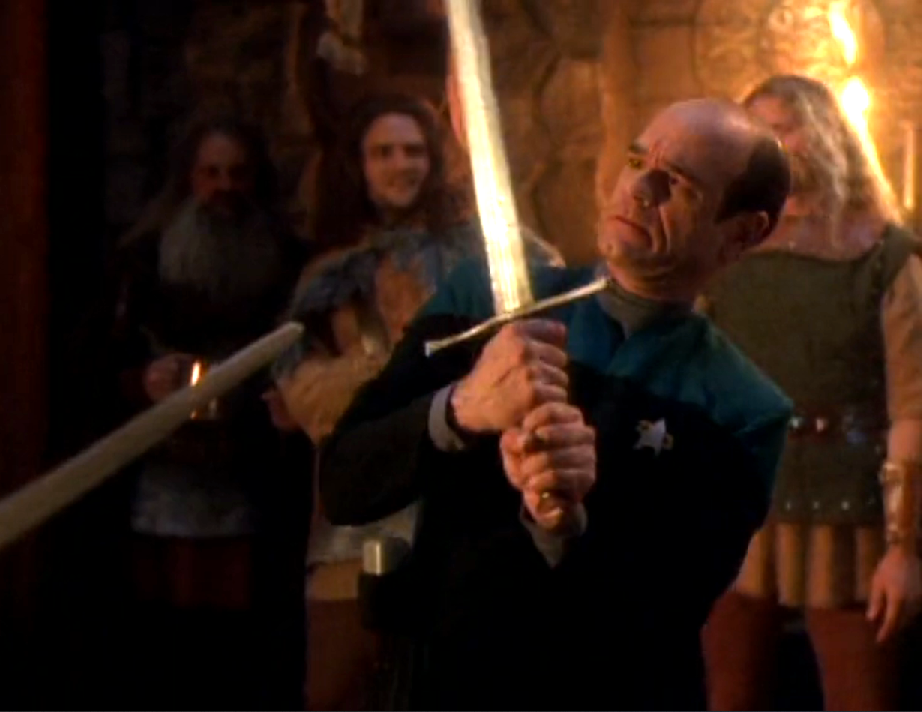 The Doctor awkwardly holding a sword while onlookers smile