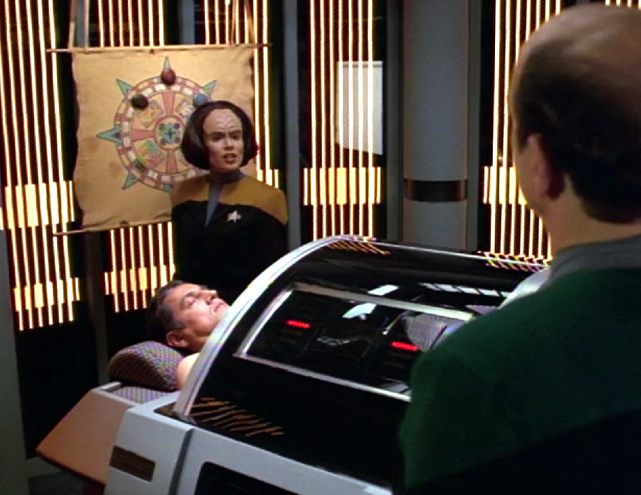 B'Elanna and the Doctor standing over Chakotay who is lying, unconscious, on a bed in sickbay