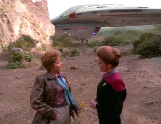 On the surface of a planet, Janeway and a woman in a leather jacket are talking in the foreground while the USS Voyager rests in the background, having landed on the surface
