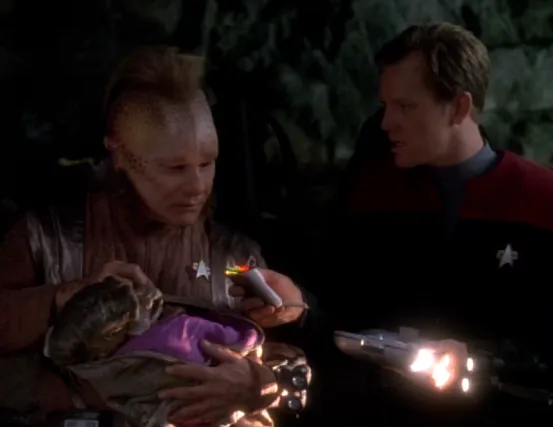 Neelix holds an alien infant while Paris scans it with his tricorder