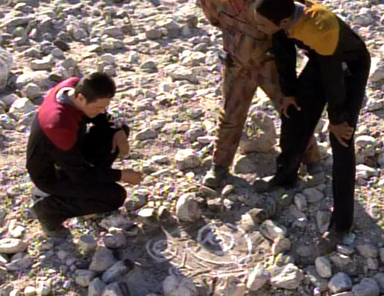 Chakotay kneels on a planet's surface to inspect a strange symbol with lines and swirls