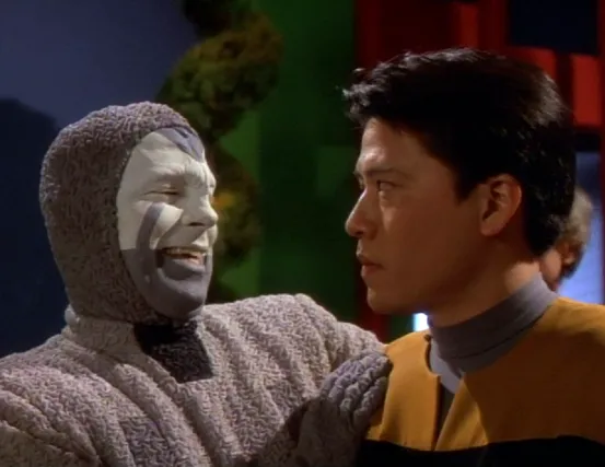 A man in a grey costume with white and grey face paint laughs as a concerned Harry Kim looks on