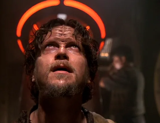 A goateed man looking upward, his head haloed by the red glow of a chute that Harry is working on in the background