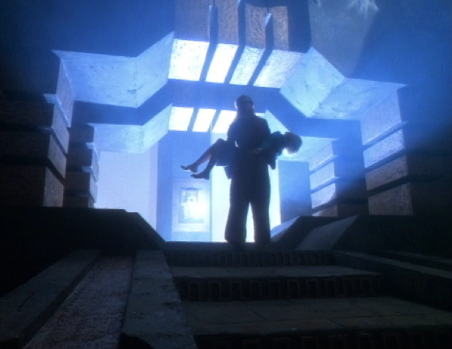Janeway, carrying an unconscious Kes, standing before a gateway with light pouring out of it