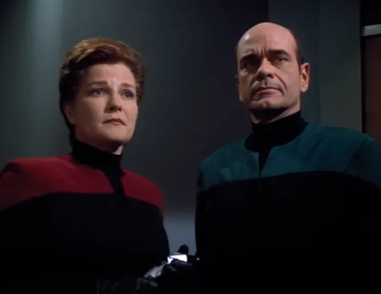 Janeway with a shorter haircut and the Doctor with yellow eyes both wearing uniforms with black gloves and black undershirts