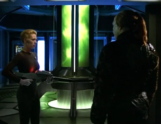 Seven carrying a phaser rifle and talking with an alien man in front of the warp core, which has turned green