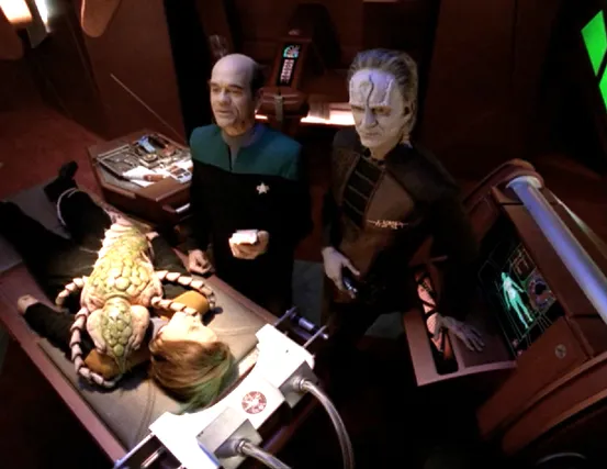 Torres lying in a medical lab with an insect-like alien attached to her as the Doctor and a Cardassian stand over her running scans