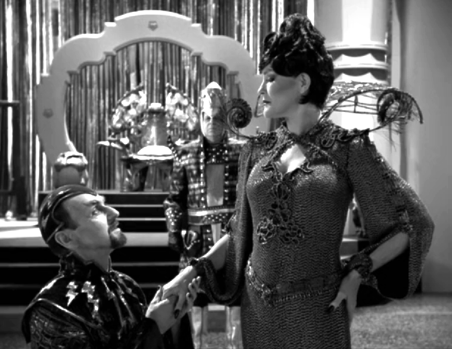 A black-and-white scene with a man kneeling before Janeway, who is wearing an elaborate dress