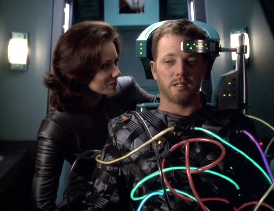 A woman speaking to Tom, who is in the pilot seat of a shuttlecraft and wearing a suit with numerous tubes connecting him to the ship