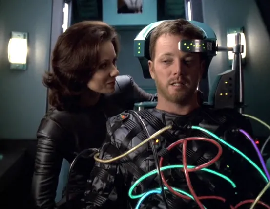 A woman speaking to Tom, who is in the pilot seat of a shuttlecraft and wearing a suit with numerous tubes connecting him to the ship