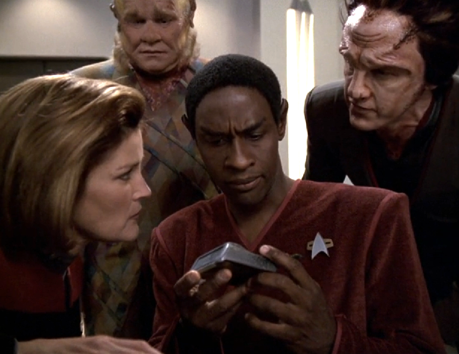 Tuvok looking quizzically at a damaged tricorder while Janeway, Neelix, and an alien man watch him with anticipation
