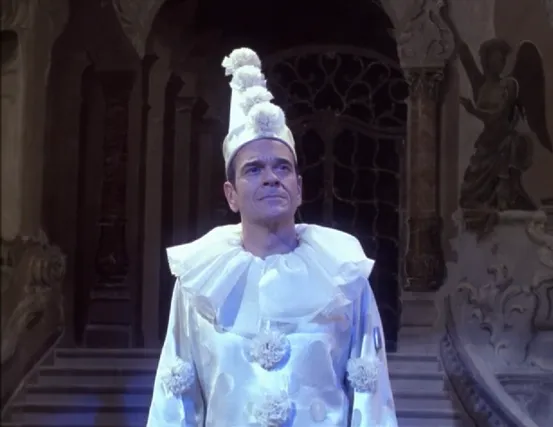 The Doctor, on a theater stage, wearing a white, polka-dotted, satin clown suit with chiffon collar and flowers down the front of the shirt and along the sleeves