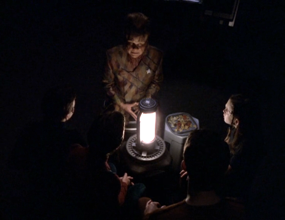 Neelix and the four Borg children in a dark room, huddled around a single battery-operated light