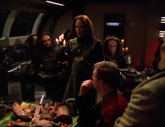 Torres, in a Klingon dress, telling a story to a room full of Klingon warriors