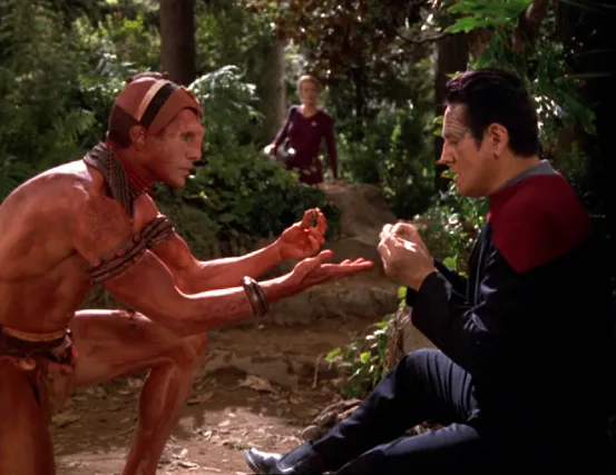 Chakotay communicating with a primitive local on a planet as Seven watches
